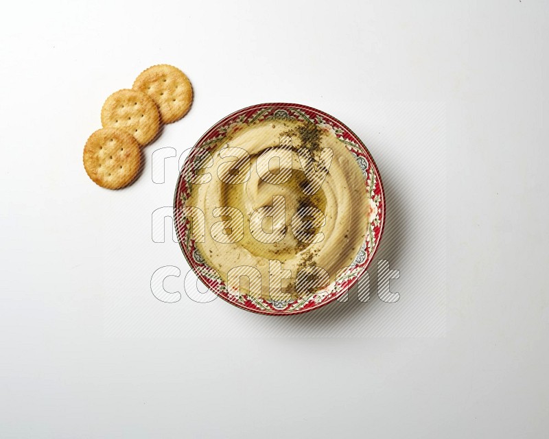 Hummus in a red plate with patterns garnished with zaatar & sumak on a white background