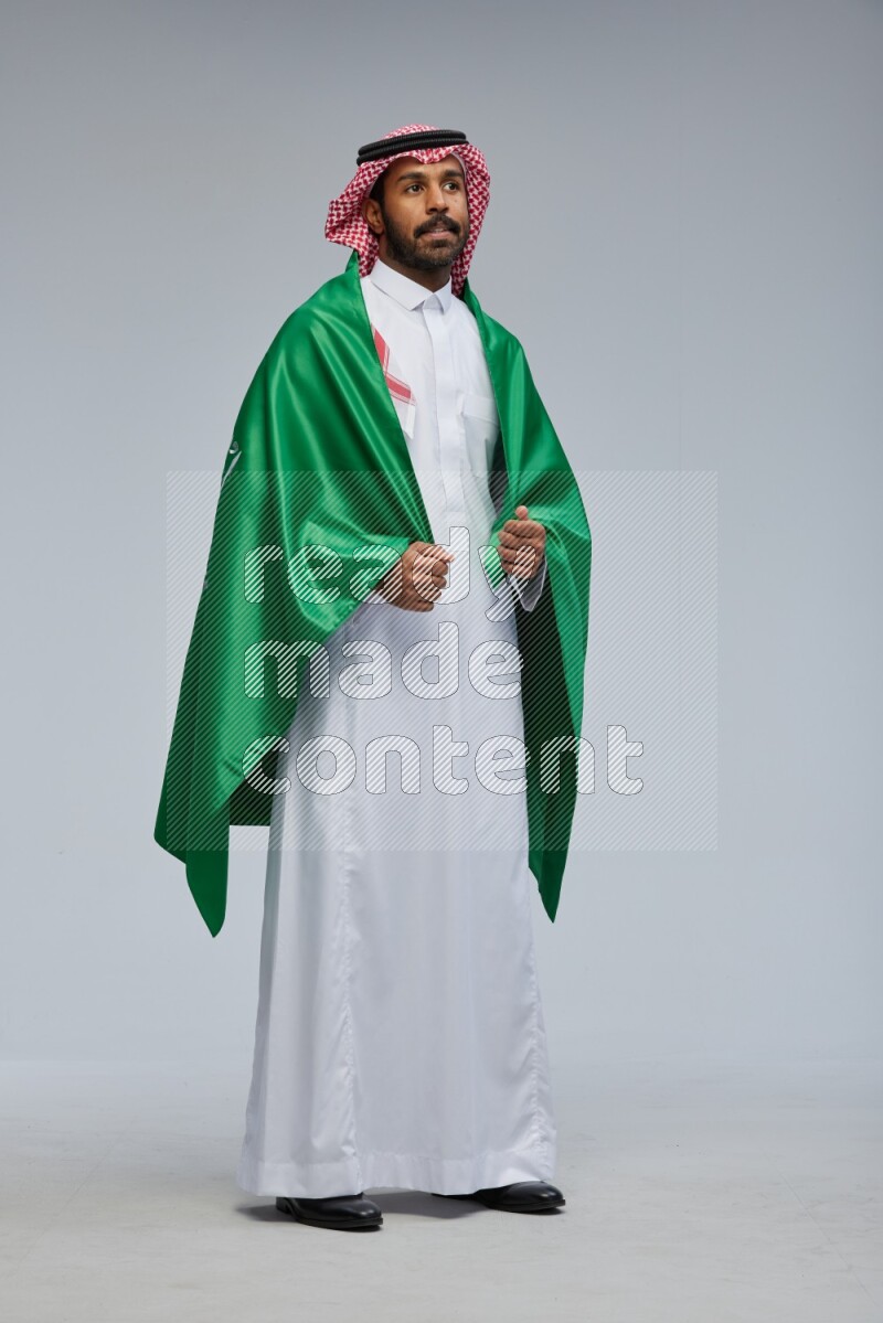 Saudi man Wearing Thob and shomag standing holding Saudi flag on Gray background