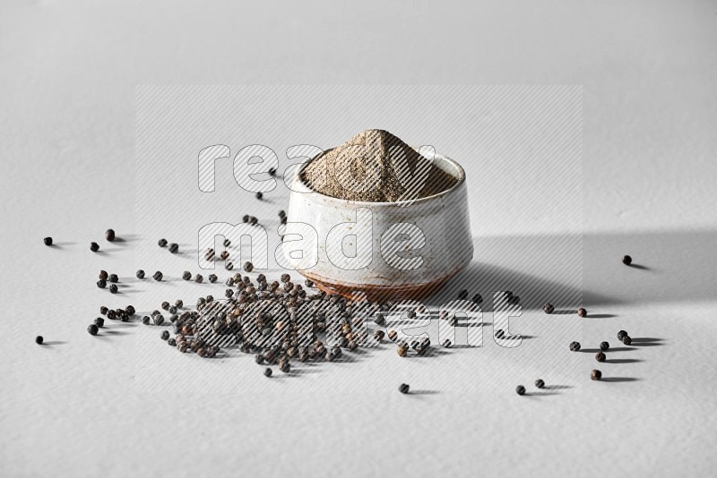 A white ceramic bowl full of black pepper powder and black pepper beads spread on white flooring