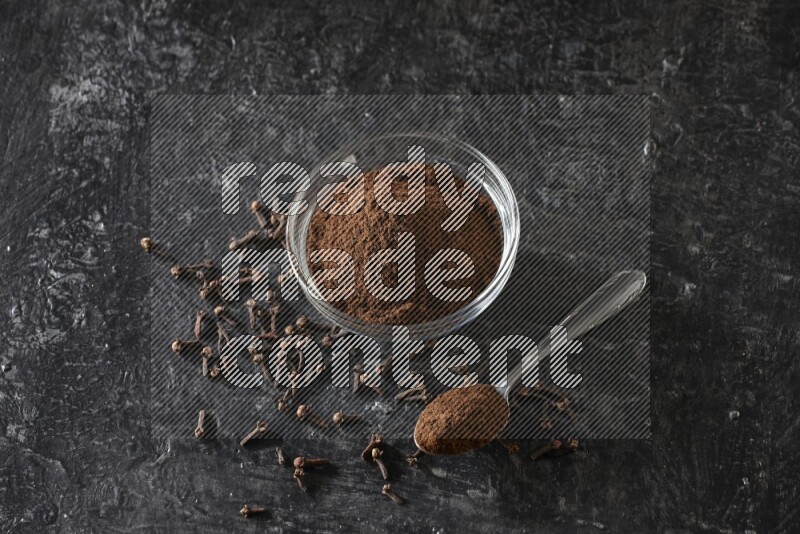 A glass bowl and a metal spoon full of cloves powder with gloves grains beside them on a textured black flooring
