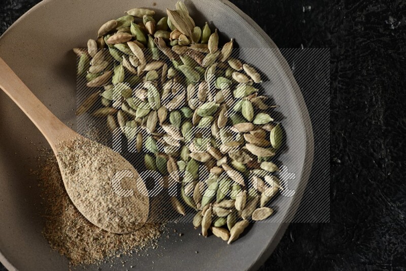 صحن ممتلئ بحبوب الهيل وملعقة خشبية ممتلئة ببودرة الهيل داخل الصحن على خلفية سوداء