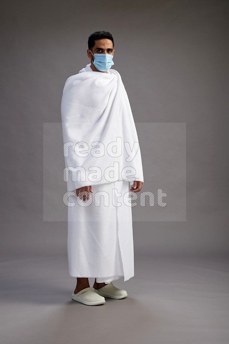 A man wearing Ehram with face mask Standing Interacting with the camera on gray background