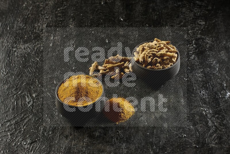 2 pottery black bowls, one full of turmeric powder and the other full of dried turmeric whole fingers on a textured black flooring