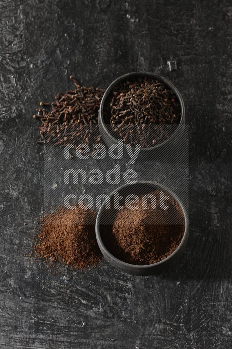 2 Black pottery bowls full of cloves and the other full of cloves powder on textured black flooring