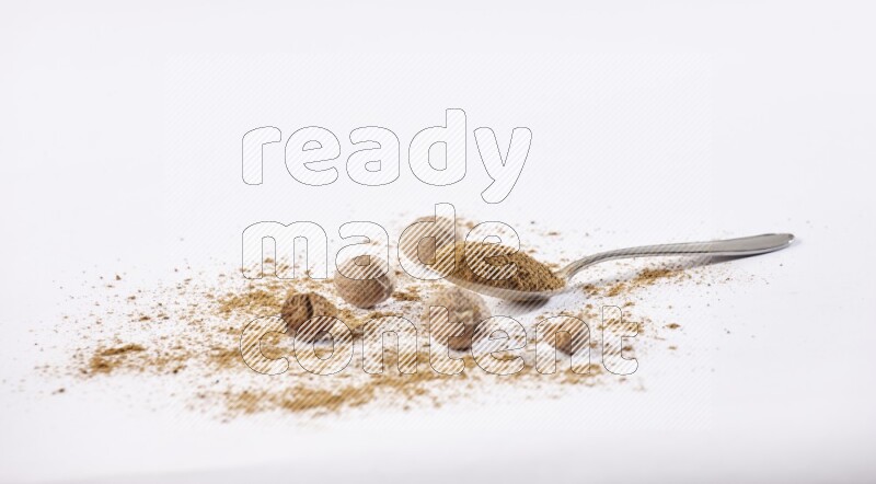 A metal spoon full of nutmeg powder with some of the powder and seeds beside it on a white flooring