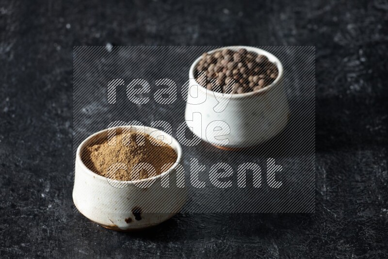 2 beige bowls, one full of allspice powder and the other full of whole balls on a textured black flooring
