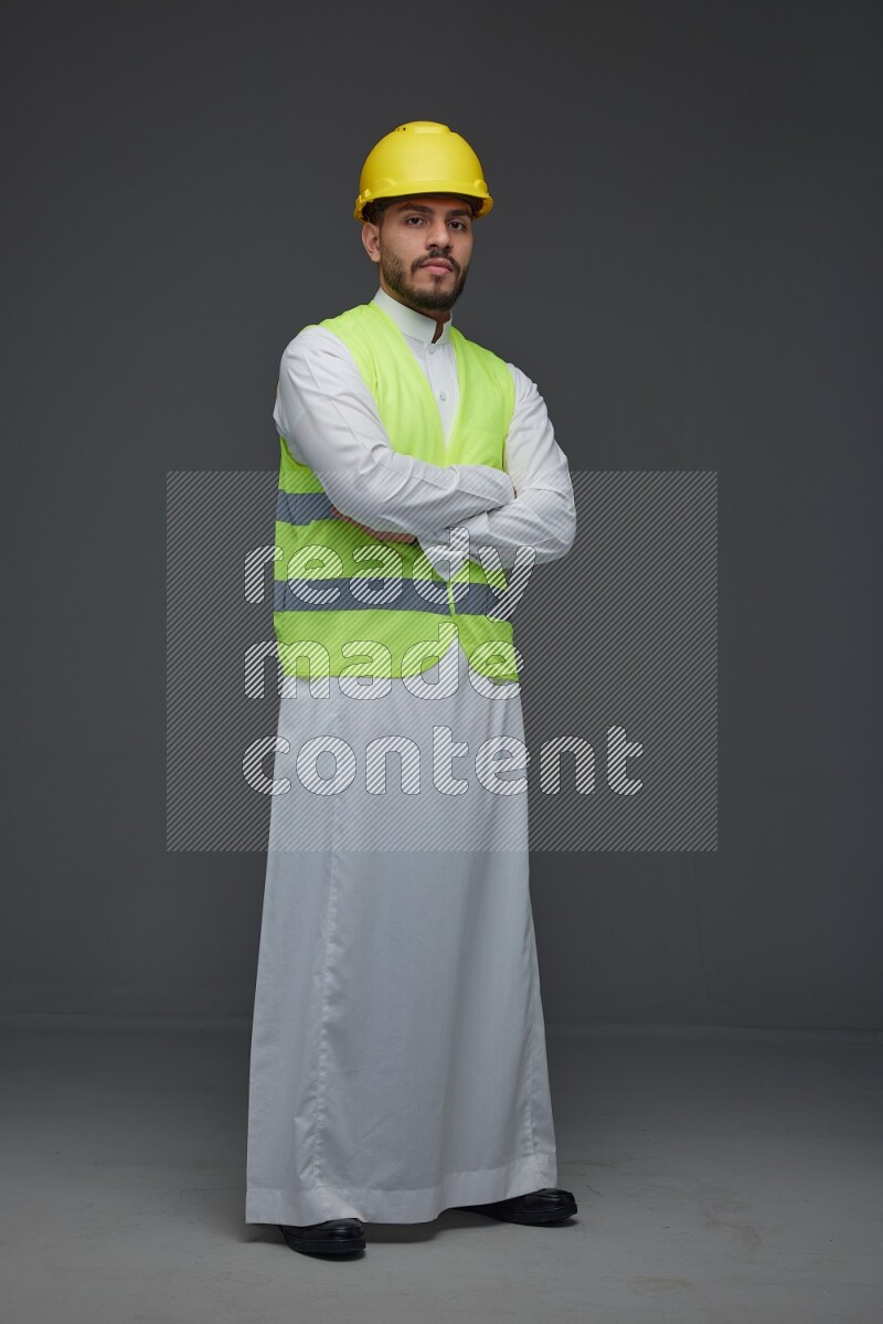 A Saudi man wearing Thobe with a yellow safety vest and white helmet standing and crossing his hands eye level on a gray background