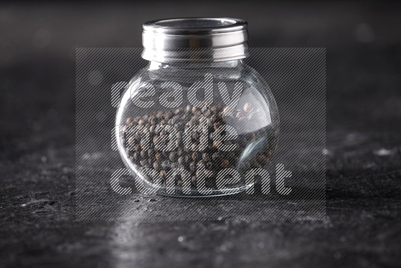A glass spice jar full of black pepper beads on textured black flooring