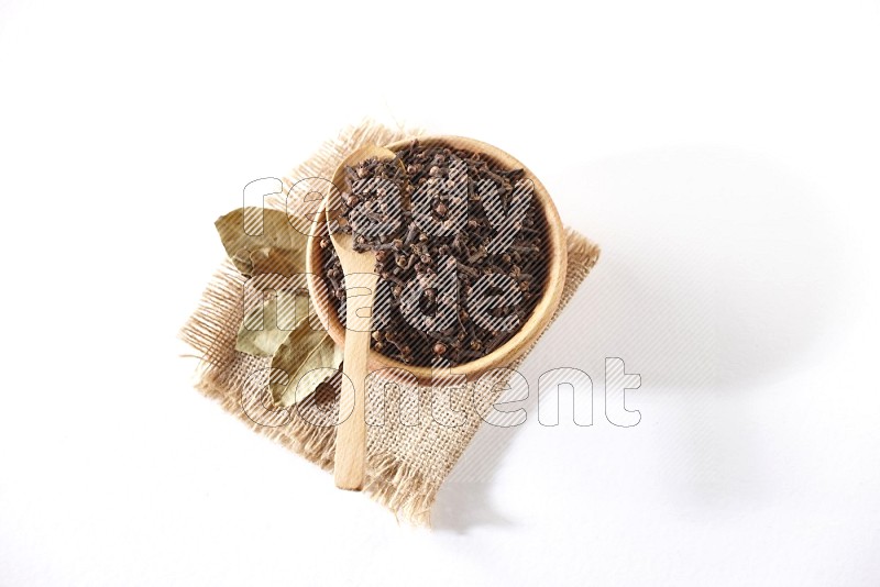 A wooden bowl and a wooden spoon full of cloves on a piece of burlap on a white flooring