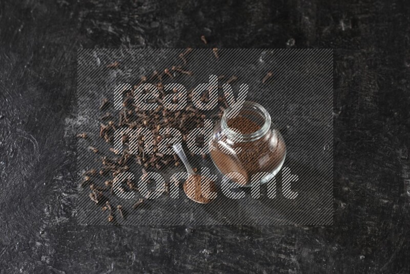 وعاء زجاجي للبهارات وملعقة معدنية ممتلئان ببودرة القرنفل مع حبوب القرنفل المتناثرة على أرضية سوداء