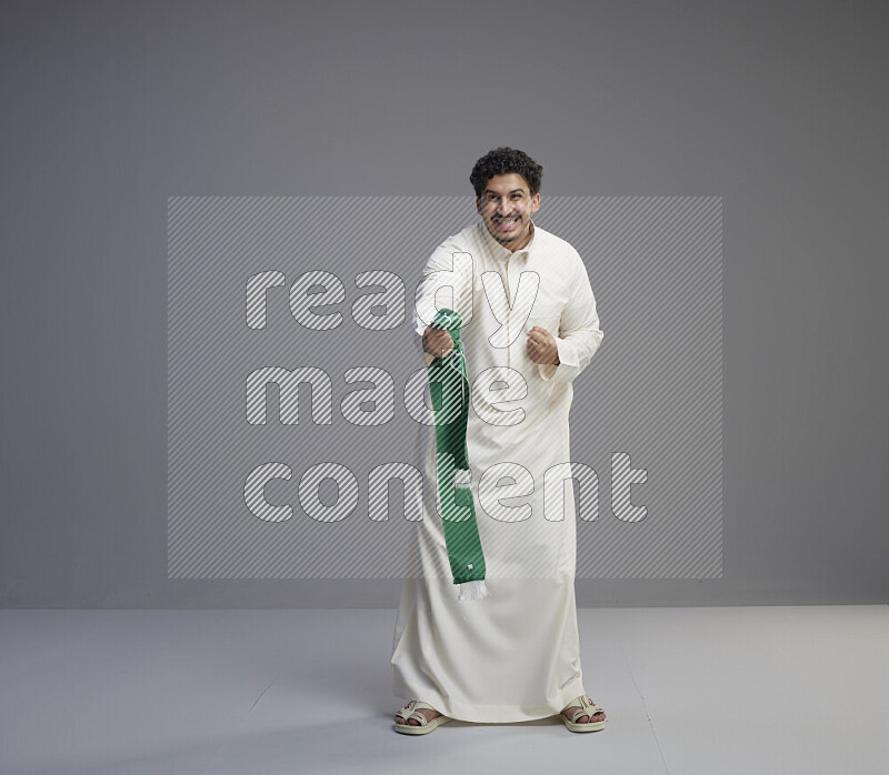 A Saudi man standing wearing thob holding Saudi flag scarf on gray background