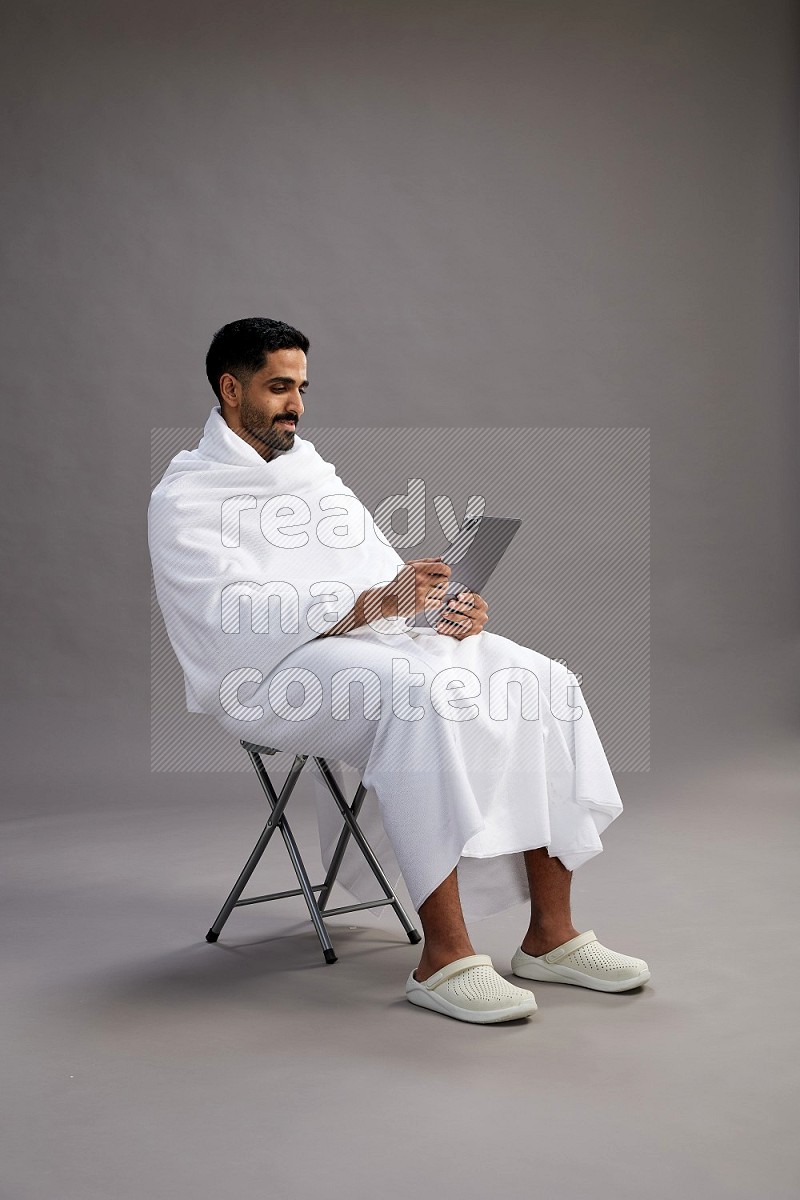 A man wearing Ehram sitting on chair holding a tablet on gray background