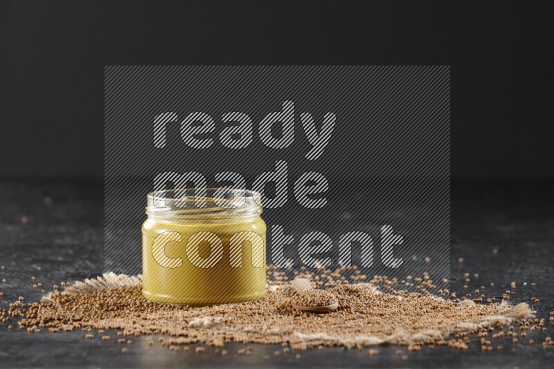 وعاء زجاجي ممتلئ بمعجون الخردل علي قطعة من القماش مع ملعقة خشبية ممتلئة بأسفله على أرضية سوداء