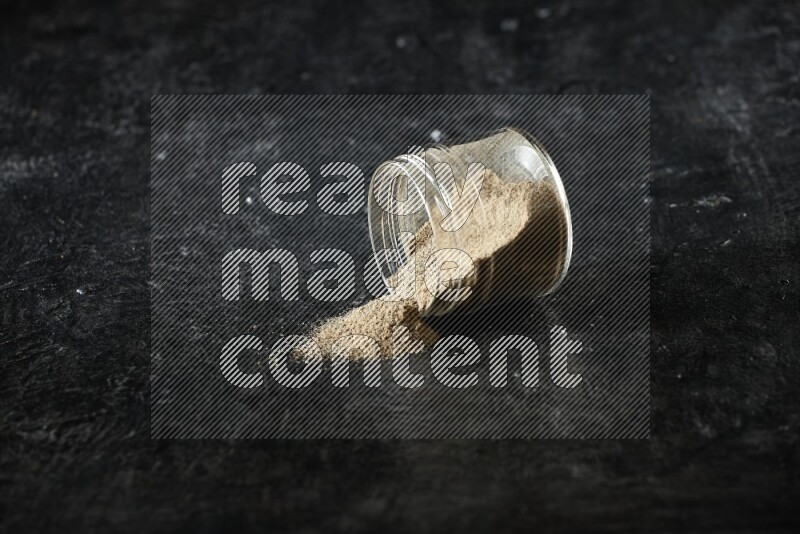 وعاء زجاجي مقلوب ممتلئ ببودرة الهيل والبودرة تناثرت منه على خلفية سوداء