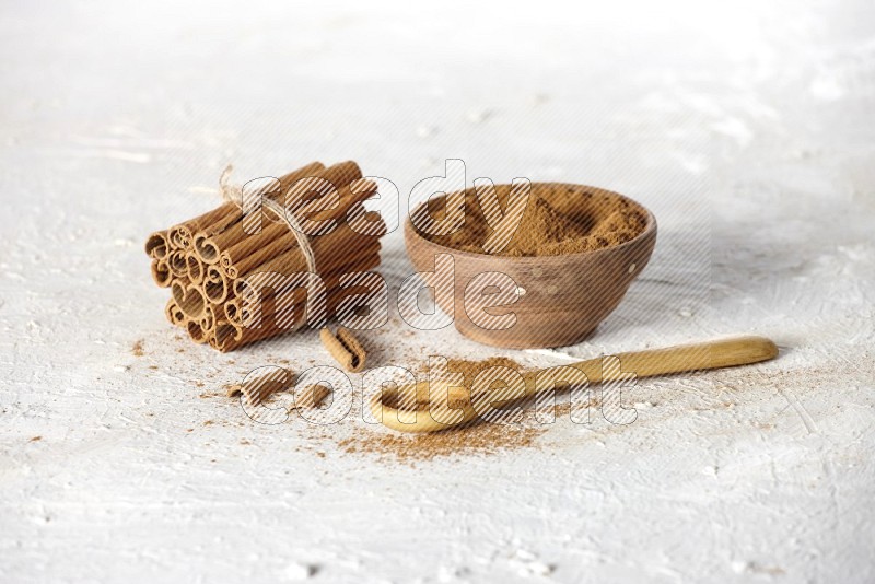 Cinnamon sticks stacked and bounded beside a wooden bowl full of cinnamon powder and a wooden spoon full of powder on white background