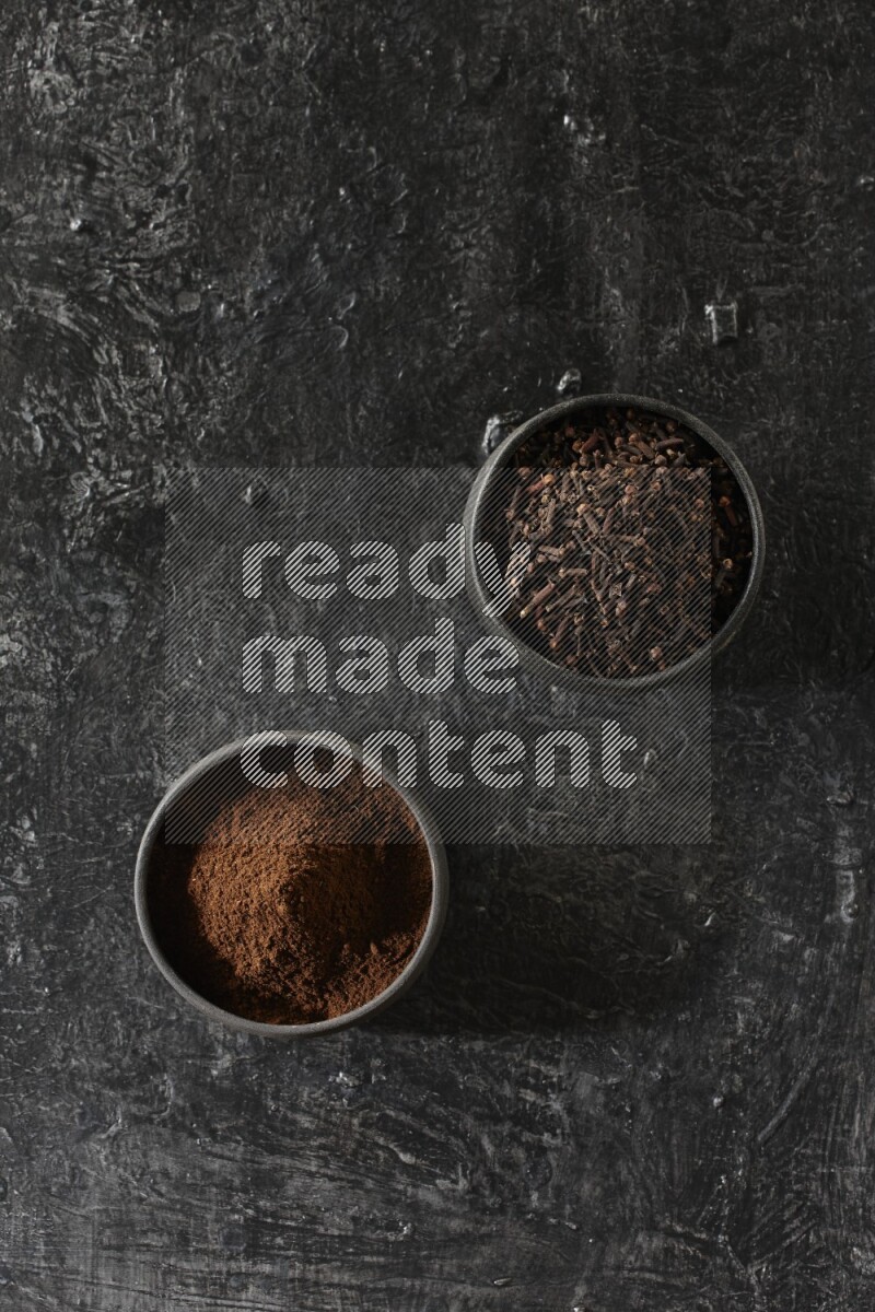 2 Black pottery bowls full of cloves and the other full of cloves powder on textured black flooring