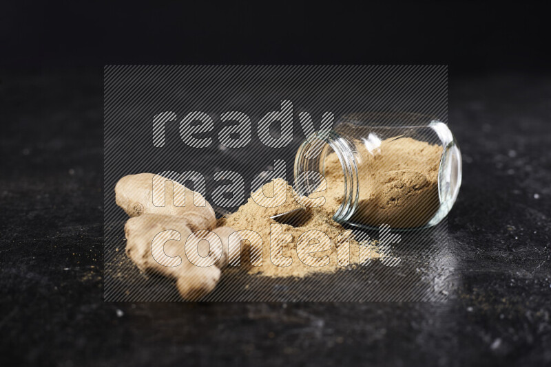 A glass jar full of ground ginger powder flipped with some spilling powder on black background