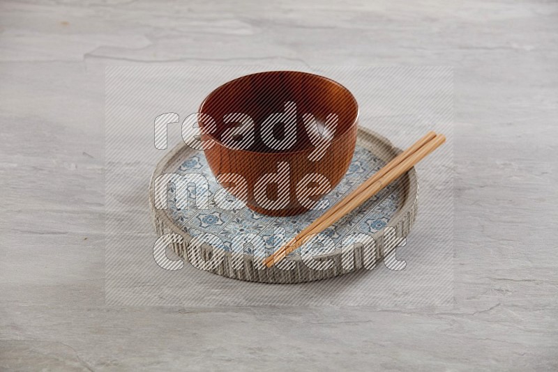 brown wood round bowl on top of multi color round ceramic plate and chopsticks, on grey textured countertop