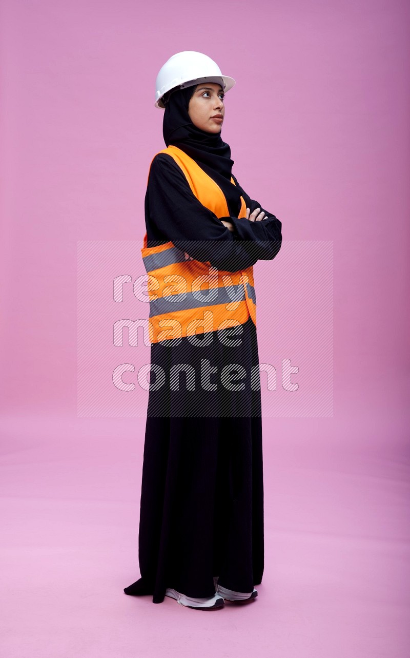 Saudi woman wearing Abaya with engineer vest and helmet standing with crossed arms on pink background