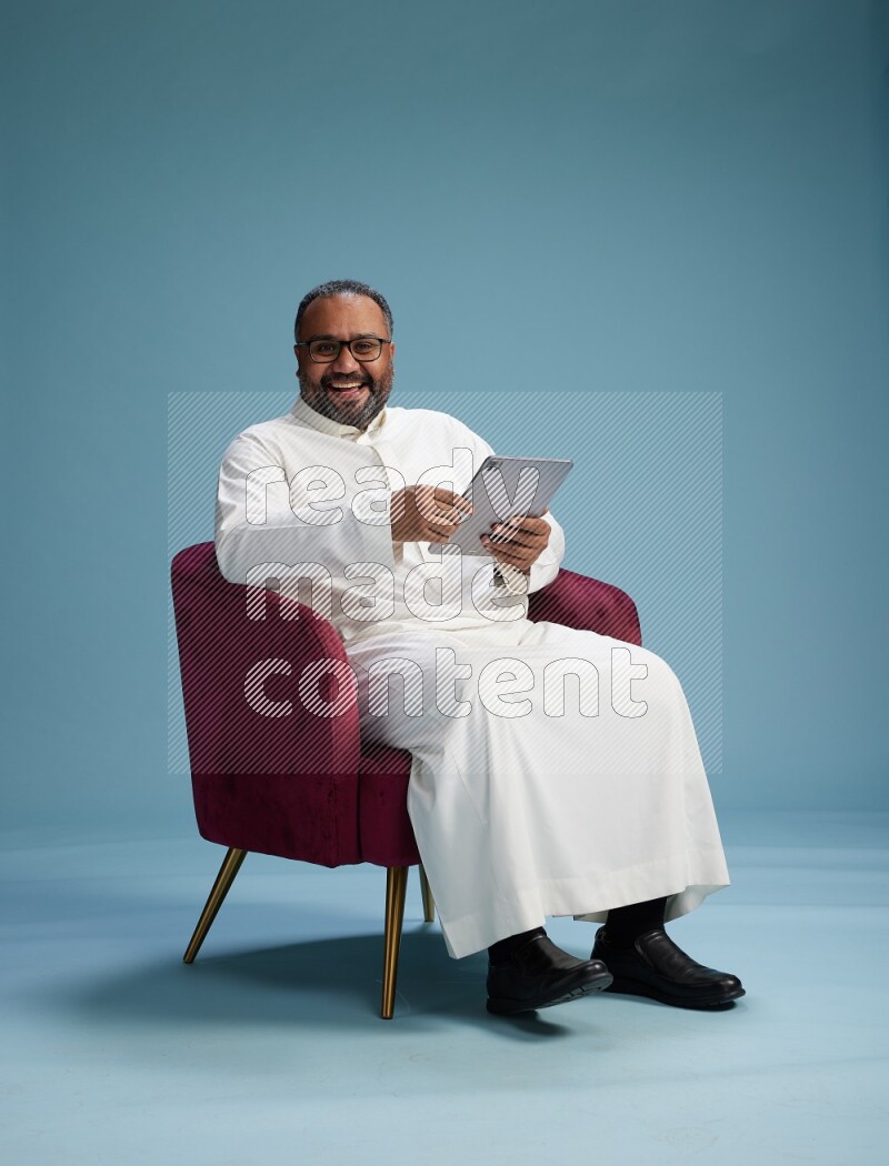 Saudi Man without shimag sitting on chair working on tablet on blue background