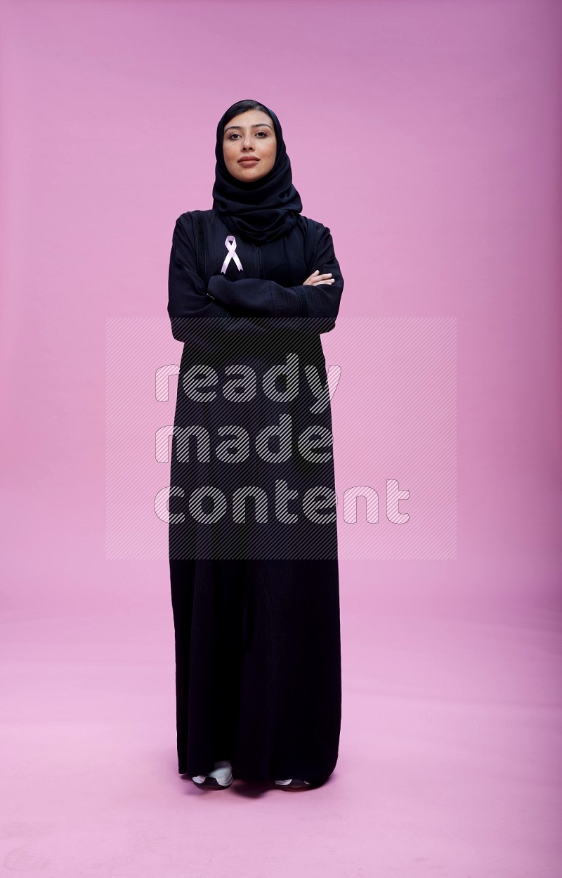 Saudi woman wearing pink ribbon on Abaya standing crossed arms on pink background