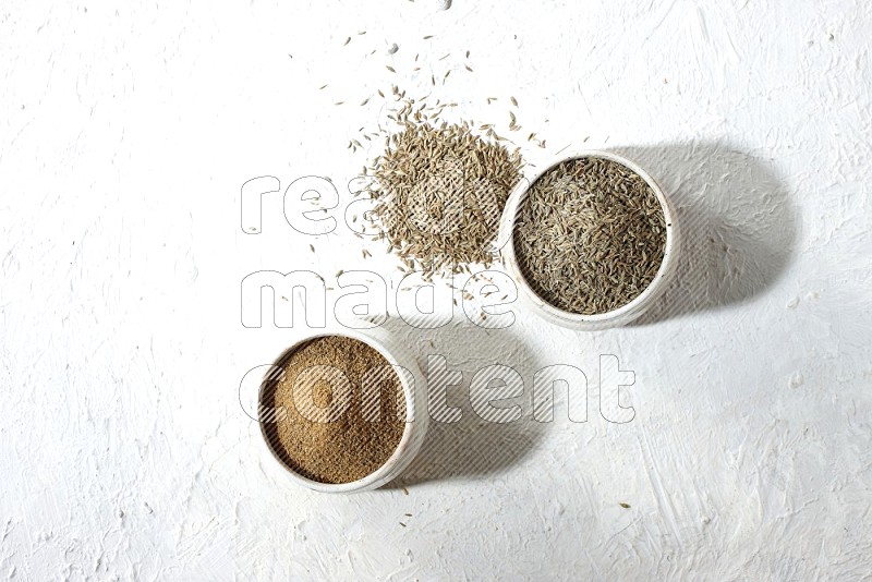 2 beige bowls full of cumin seeds and powder with spilled powder and seeds on textured white flooring