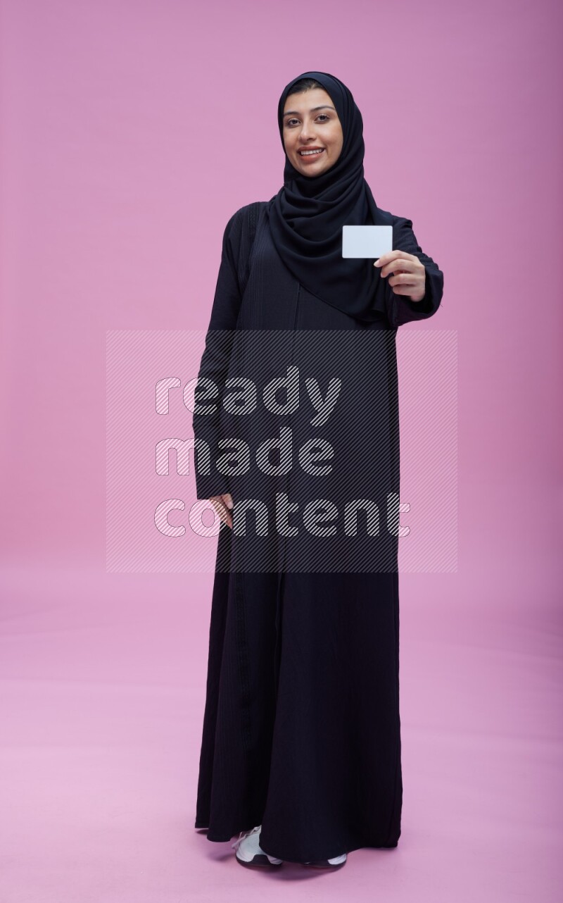 Saudi woman wearing Abaya standing holding ATM card on pink background