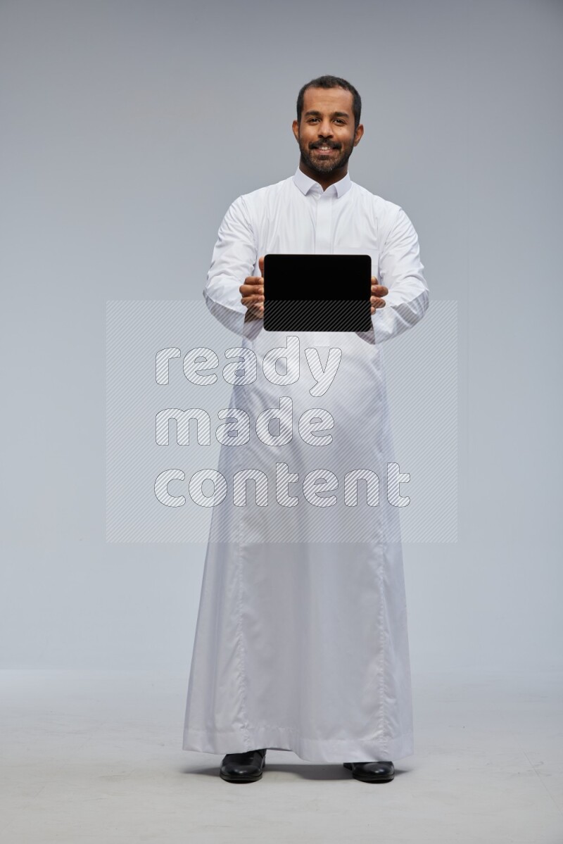 Saudi man wearing Thob standing showing tablet to camera on Gray background