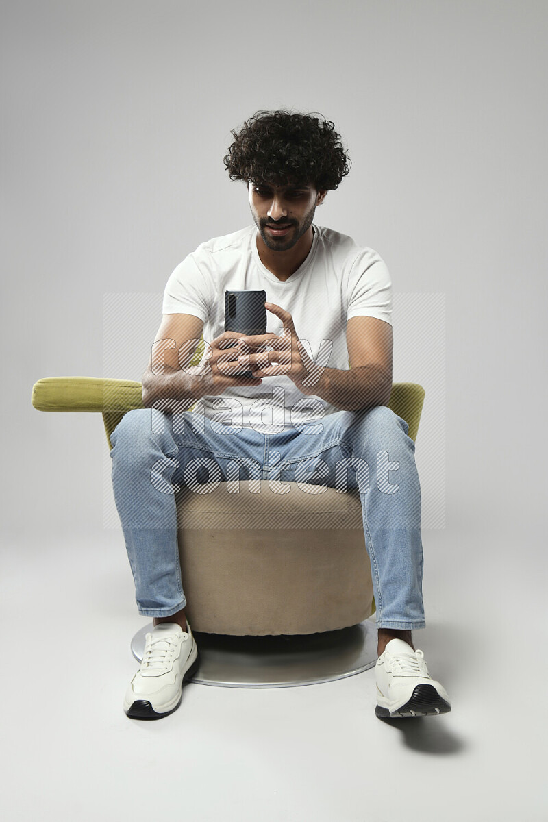 رجل يرتدي ملابس كاجوال يجلس علي كرسي و يصور بهاتفه علي خلفية بيضاء