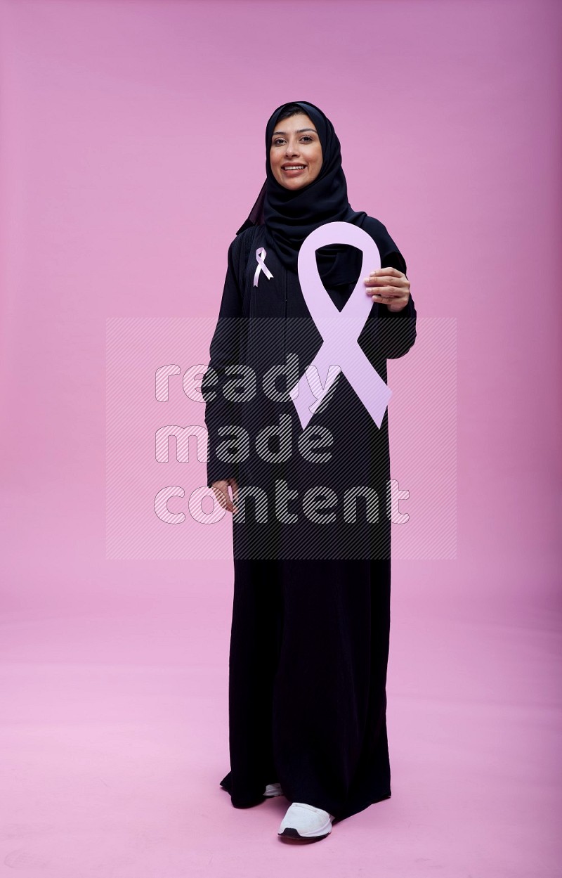 Saudi woman wearing pink ribbon on abaya standing holding awareness ribbon on plain pink background