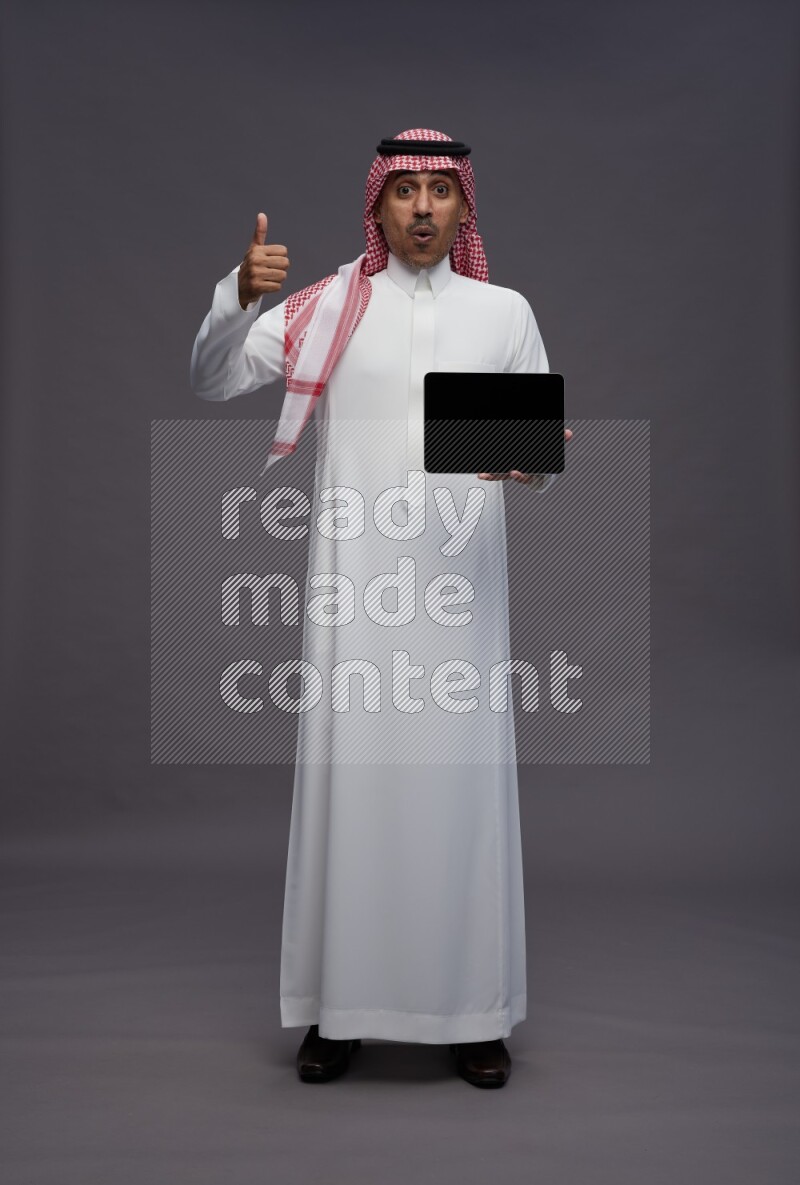 Saudi man wearing thob and shomag standing showing tablet to camera on gray background