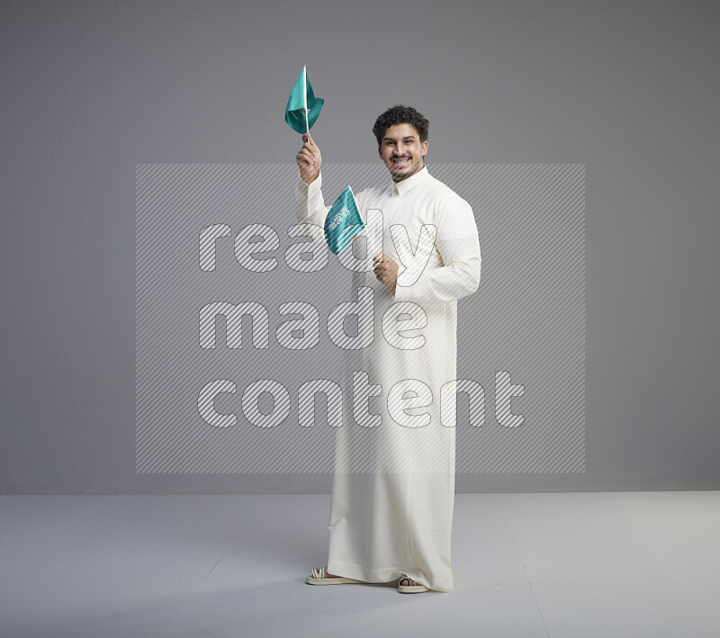 A Saudi man standing wearing thob holding small Saudi flag on gray background