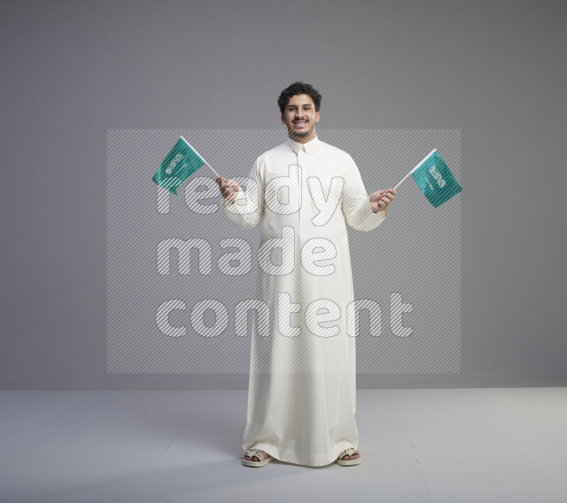 A Saudi man standing wearing thob holding small Saudi flag on gray background