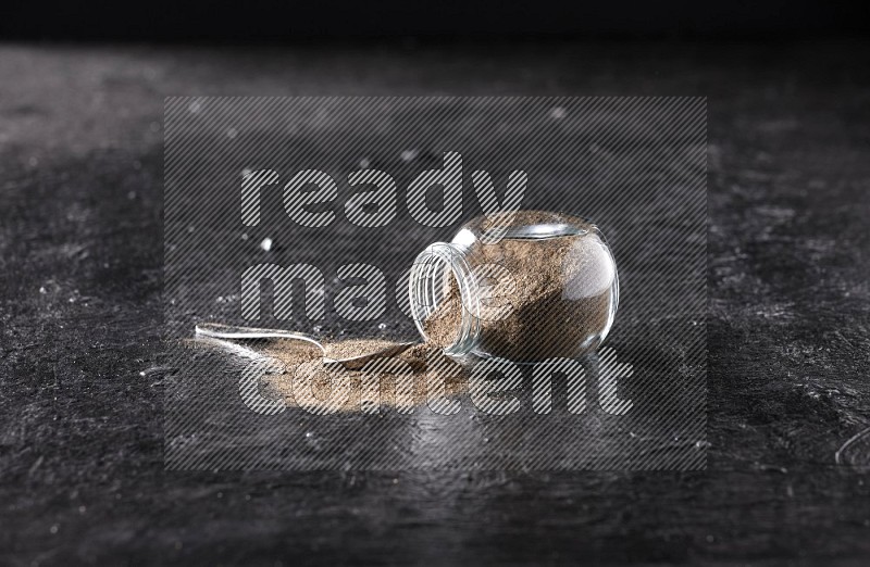 A flipped glass spice jar full of black pepper powder with a metal spoon full of powder on textured black flooring