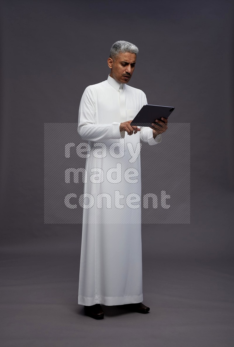 Saudi man wearing thob standing working on tablet on gray background