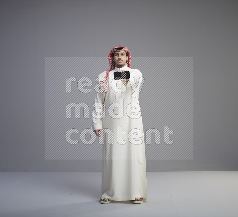 A Saudi man standing wearing thob and red shomag showing phone to camera on gray background