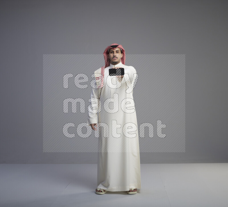 A Saudi man standing wearing thob and red shomag showing phone to camera on gray background