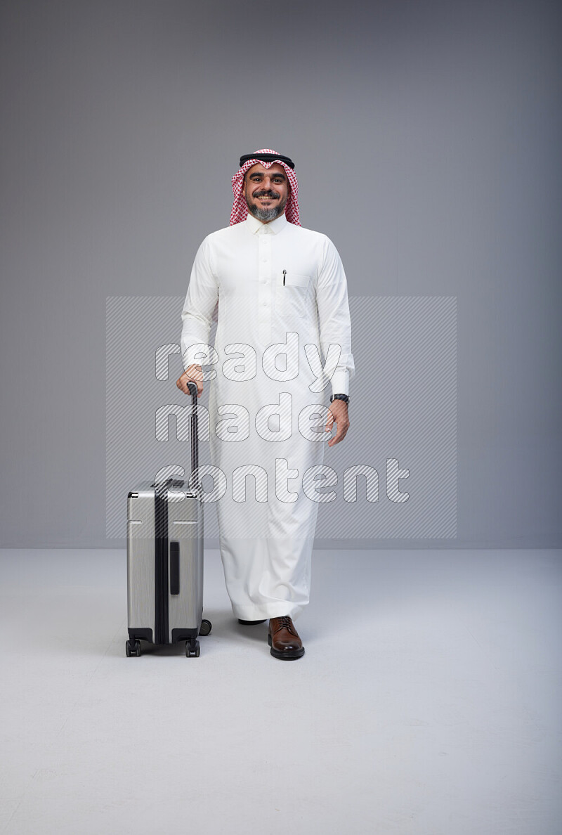 Saudi man wearing Thob and red Shomag standing holding Travel bag on Gray background