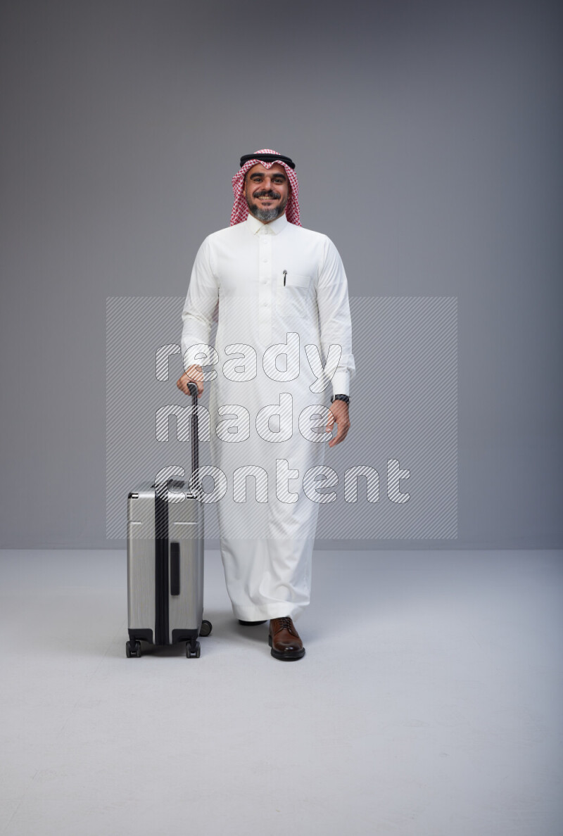 Saudi man wearing Thob and red Shomag standing holding Travel bag on Gray background