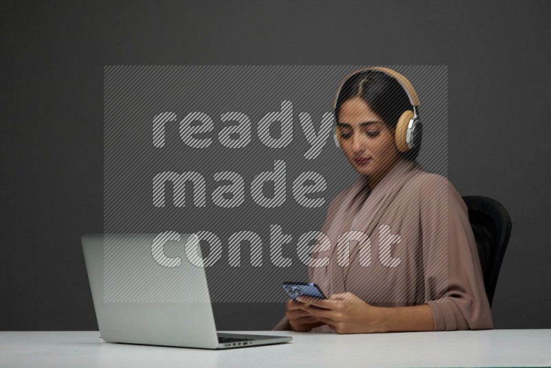 A Saudi woman Sitting on her desk Typing on her laptop wearing a headset  on a Gray Background wearing Brown Abaya