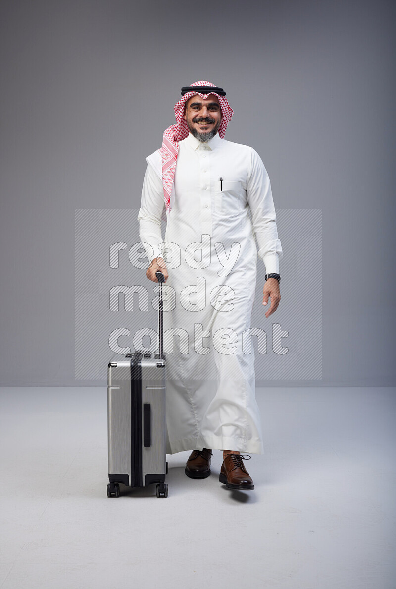 Saudi man wearing Thob and red Shomag standing holding Travel bag on Gray background