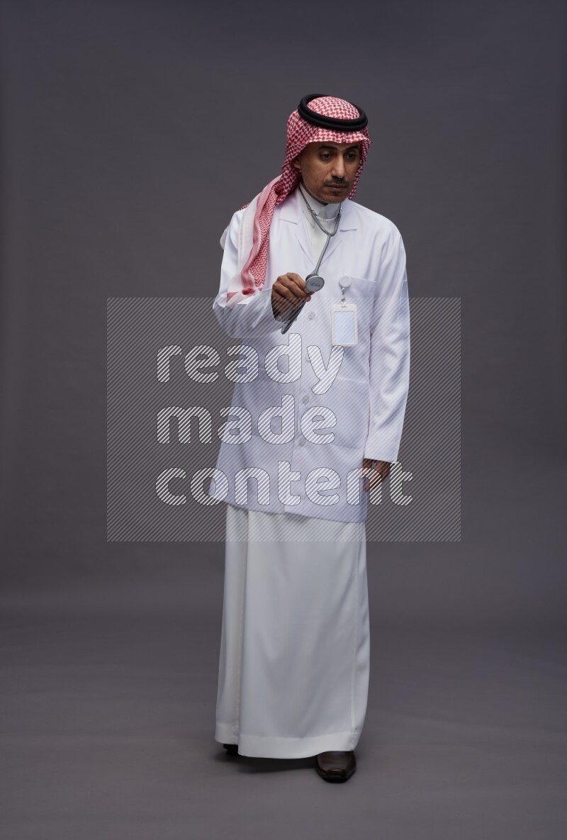 Saudi man wearing thob with lab coat and shomag with pocket employee badge with stethoscope standing interacting with the camera on gray background