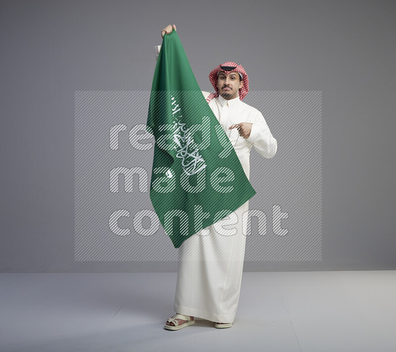 A saudi man standing wearing thob and red shomag holding big saudi flag on gray background