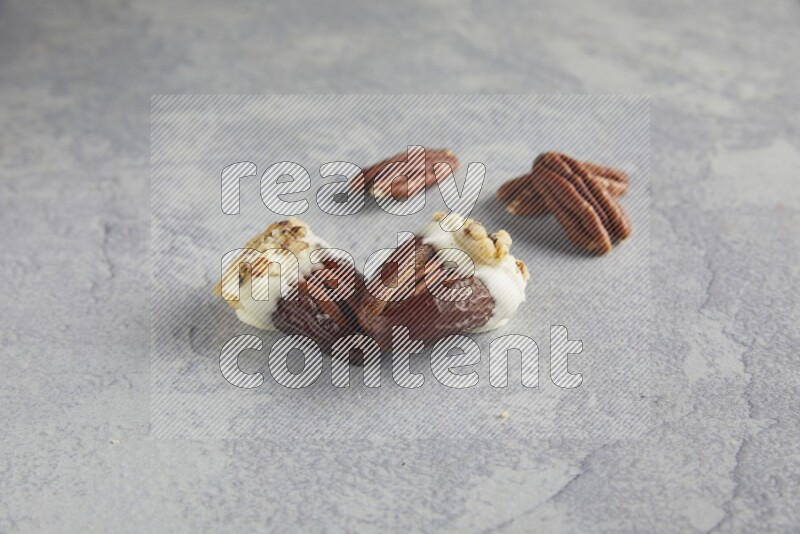 Two pecan stuffed dates covered with white chocolate and chopped walnuts with unroasted pecans on alight grey background