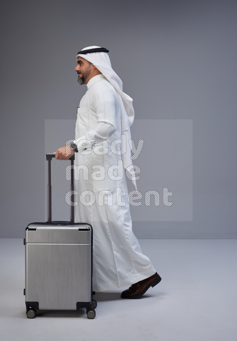 Saudi man wearing Thob and white Shomag standing holding Travel bag on Gray background