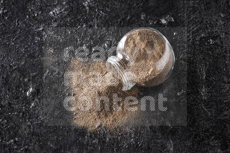 A flipped glass spice jar full of black pepper powder on textured black flooring