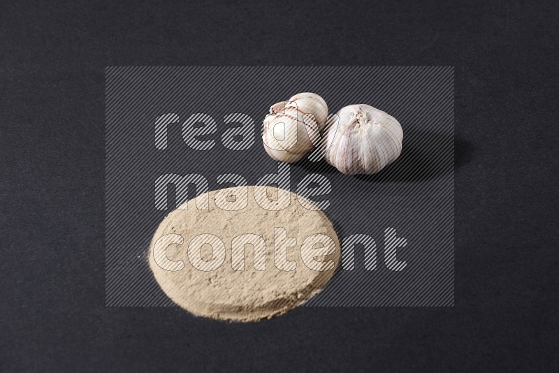 Garlic powder in a circle shape and beside it 2 garlic bulbs on a black flooring in different angles