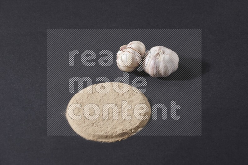 Garlic powder in a circle shape and beside it 2 garlic bulbs on a black flooring