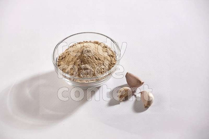 A glass bowl full of garlic powder with garlic bulb and some cloves beside it on a white flooring