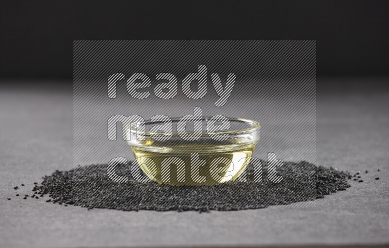 A glass bowl full of black seeds oil surrounded by the seeds on a black flooring